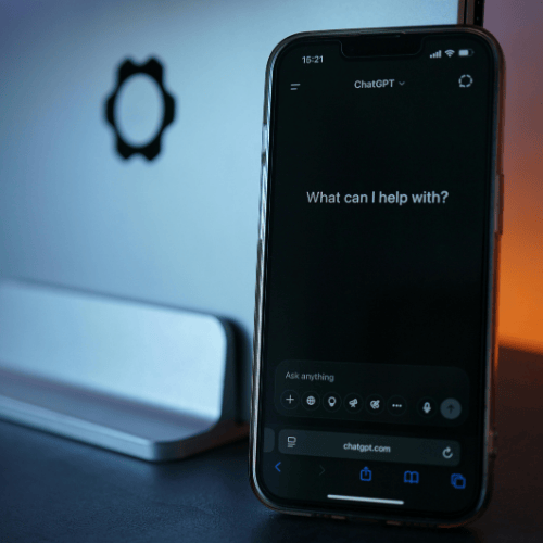 Smartphone displaying a voice assistant screen with the text “What can I help with?” placed in front of a desktop computer.