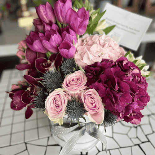 Elegant floral arrangement with pink roses, purple hydrangeas, tulips, and orchids in a white vase tied with a gray ribbon.