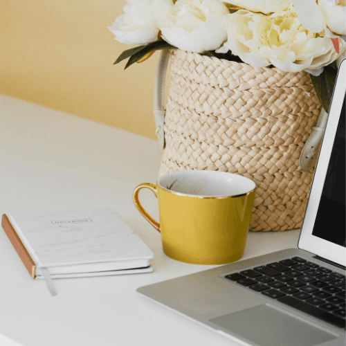 Workspace with a laptop, yellow coffee mug, notebook, and a woven basket of white flowers.
