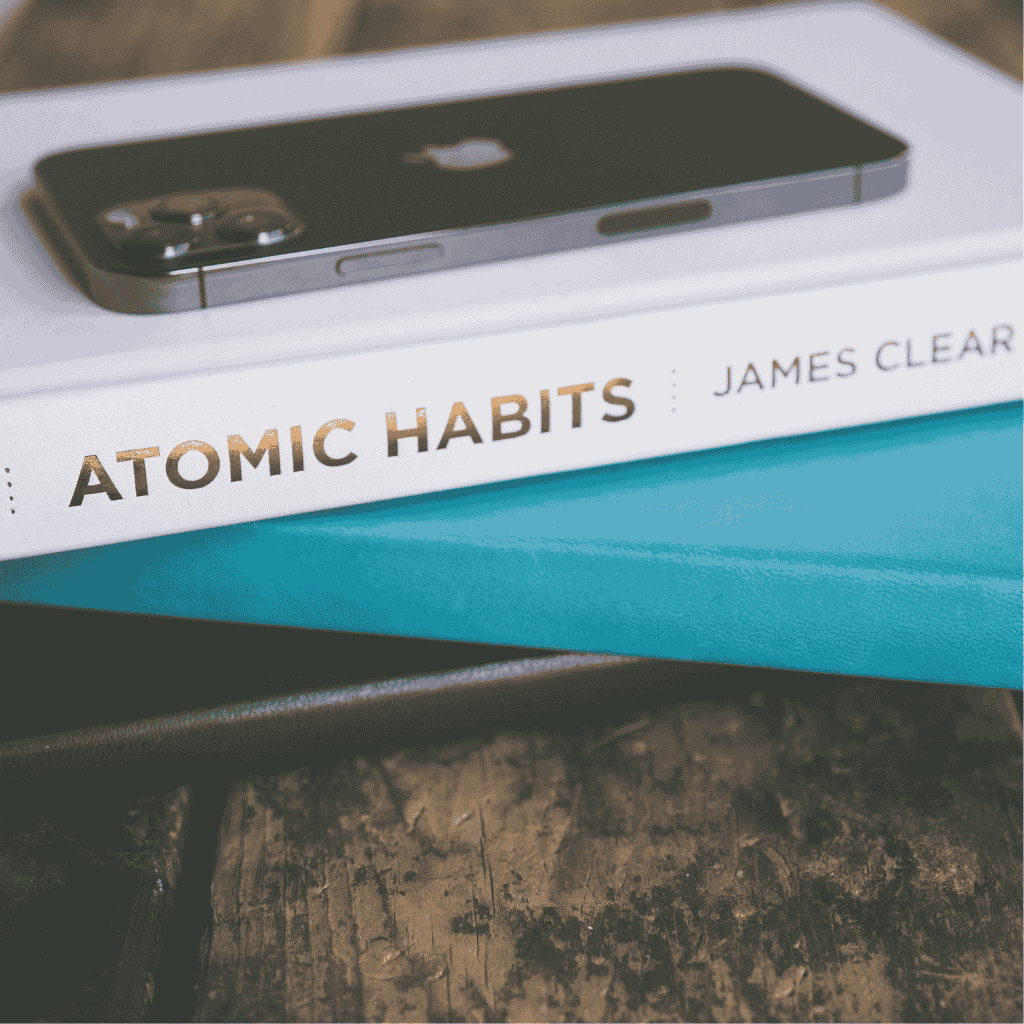 A copy of the book Atomic Habits by James Clear with an iPhone placed on top, resting on a stack of notebooks.
