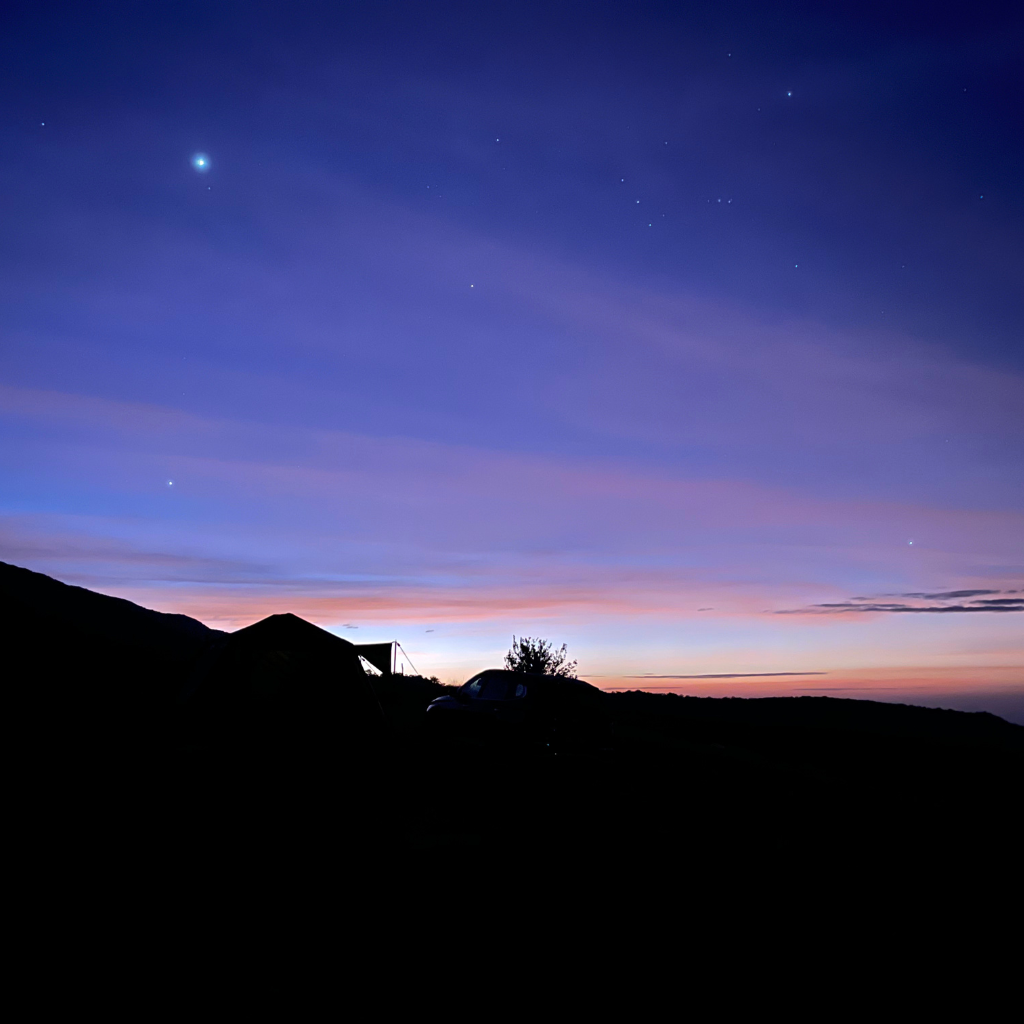 Silhouette of a tent and car under a twilight sky with stars and a bright planet visible. The horizon glows with hues of purple, blue, and orange.