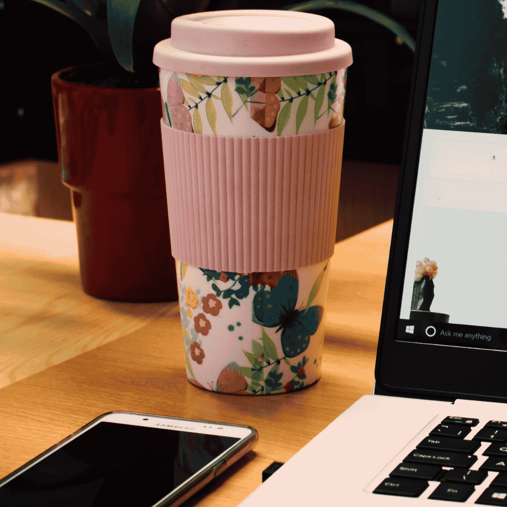 A floral reusable coffee cup with a pink silicone sleeve sits beside a laptop and smartphone on a wooden desk — a cozy setup for listening to business podcasts for entrepreneurs.