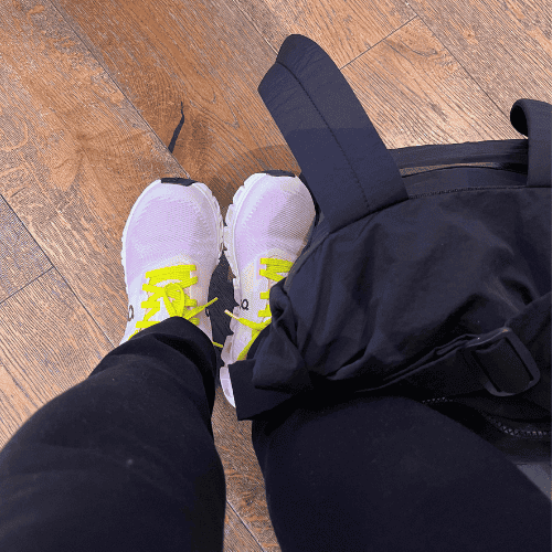 Letting go as a mother while waiting at the airport, wearing white sneakers with yellow laces beside a black travel bag.